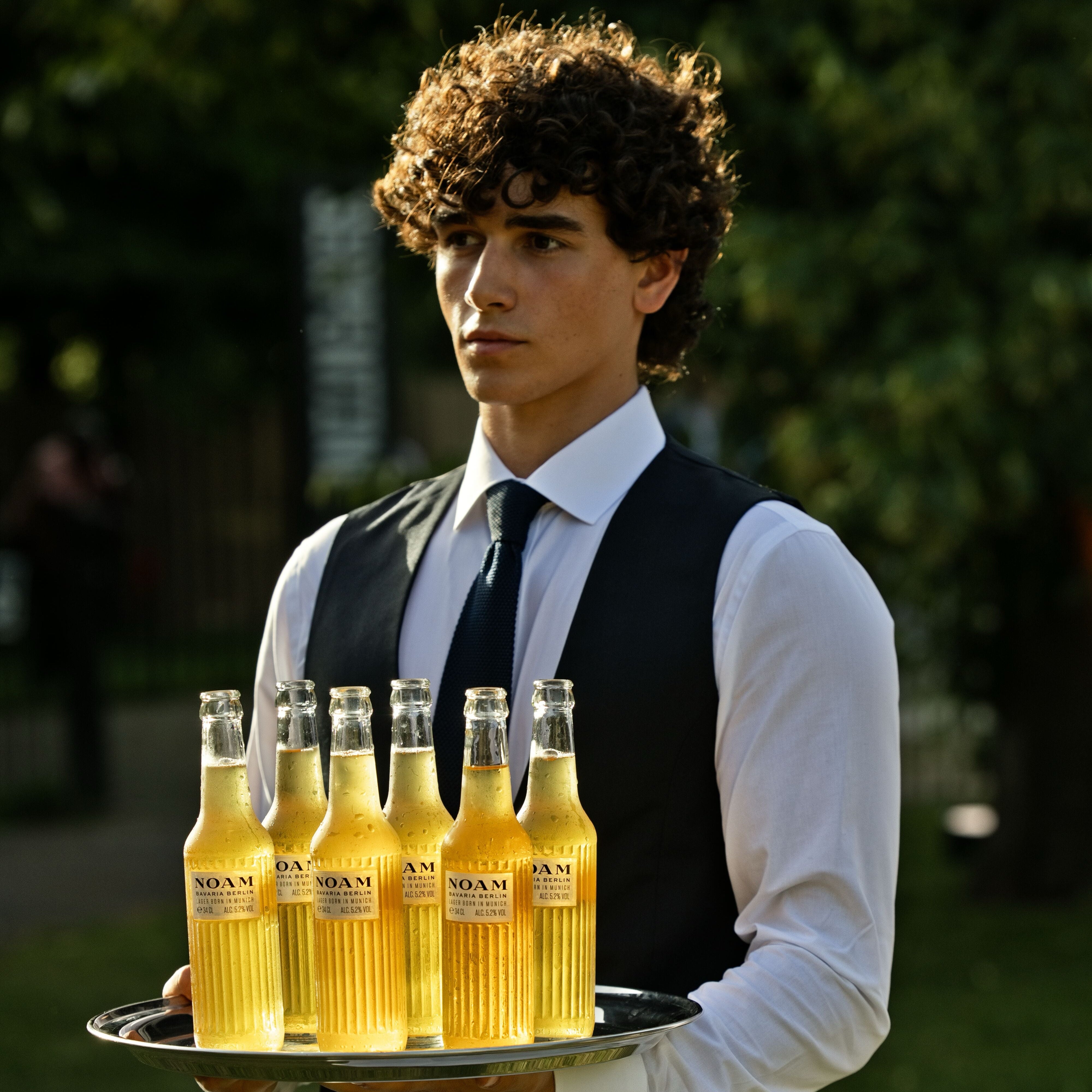 Man in formal attire holding a tray with bottles of NOAM beer outdoors