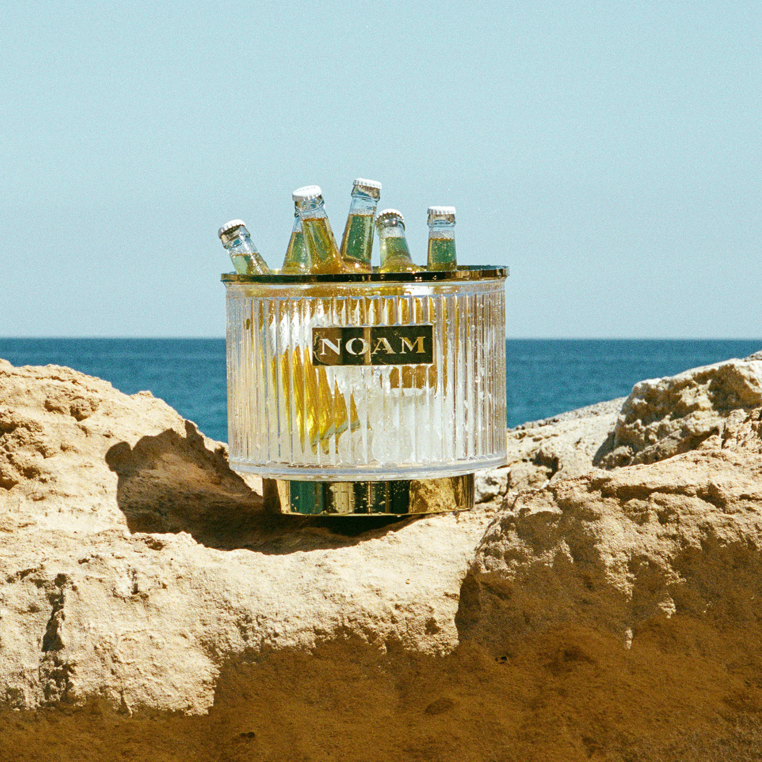 Clear ice bucket with bottles labeled 'NOAM' on a rocky surface with ocean in the background