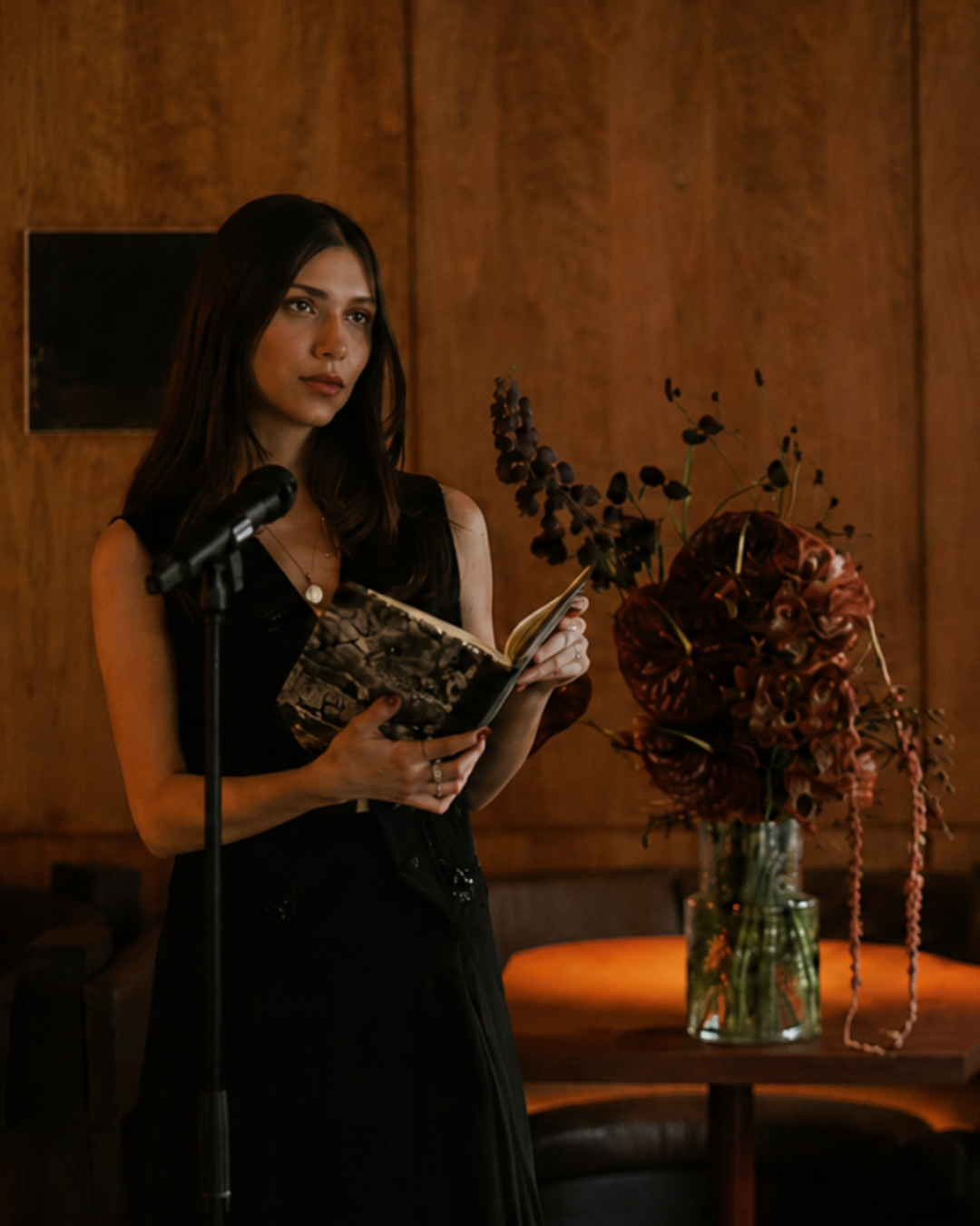 A woman in a black sleeveless dress stands at a microphone holding an open book, set against a warm wood-paneled background with a large dried floral arrangement on a table.