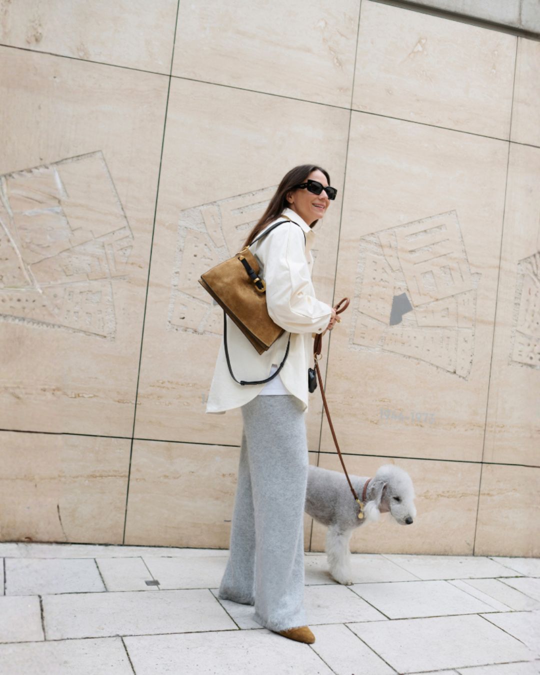 A woman in a white jacket and light gray pants walks a small gray dog along a paved street next to a beige stone wall with geometric carvings.