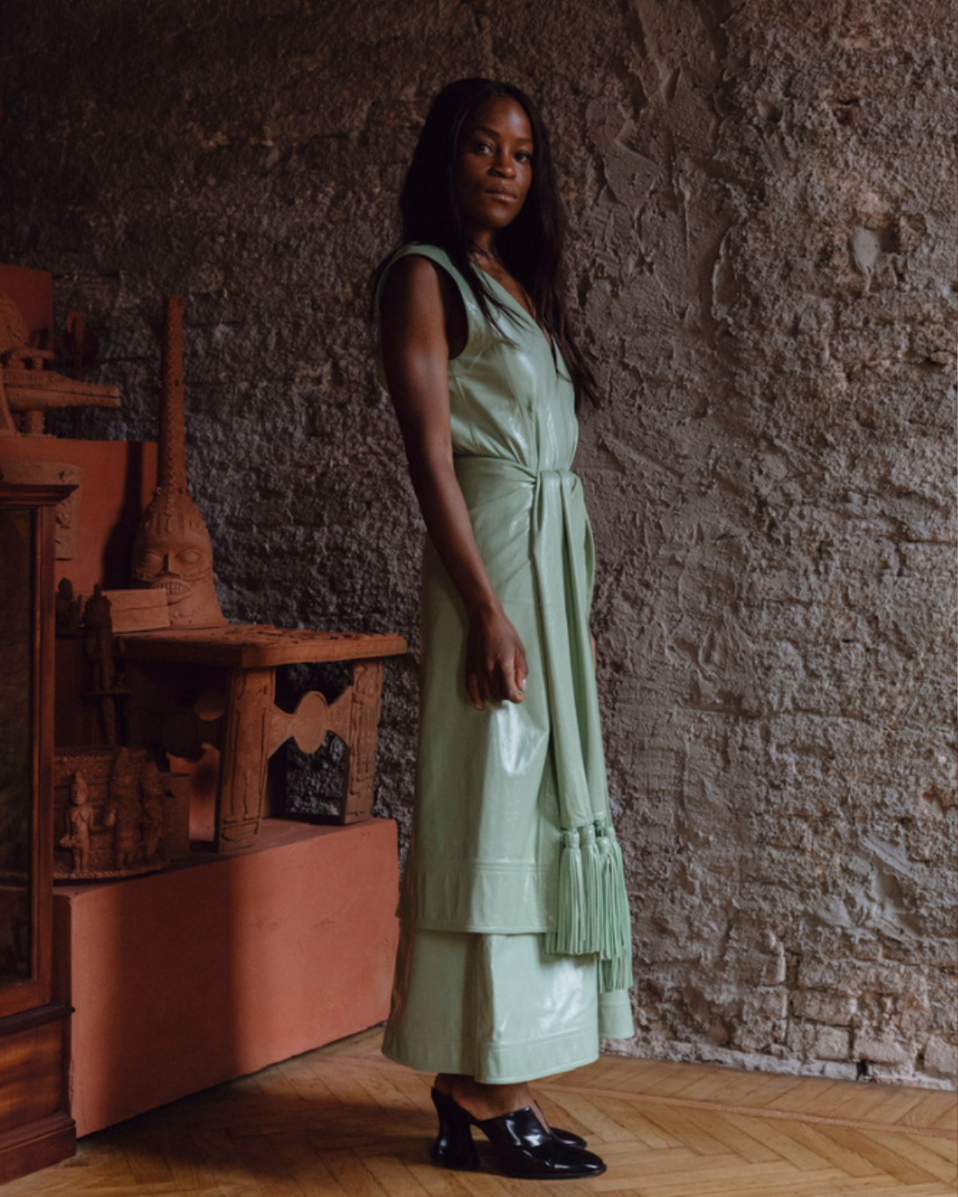 Woman in a light green dress standing against a textured wall with wooden furniture.