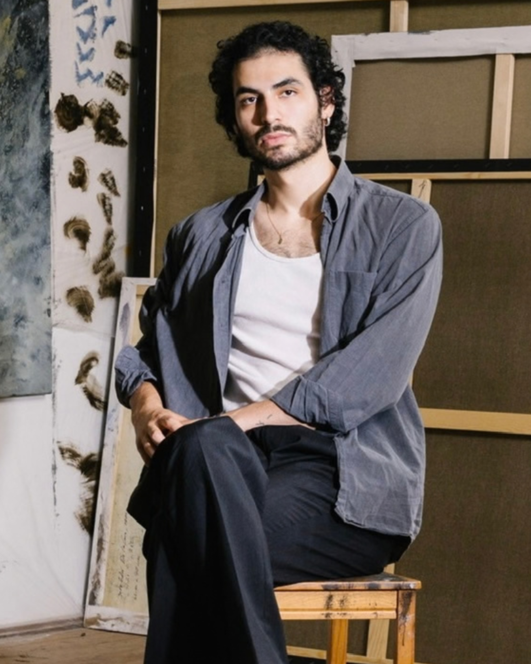 A man sits in a painterly studio interior on the left, with framed canvases leaning against the wall and a large abstract artwork hanging on the right wall. 
