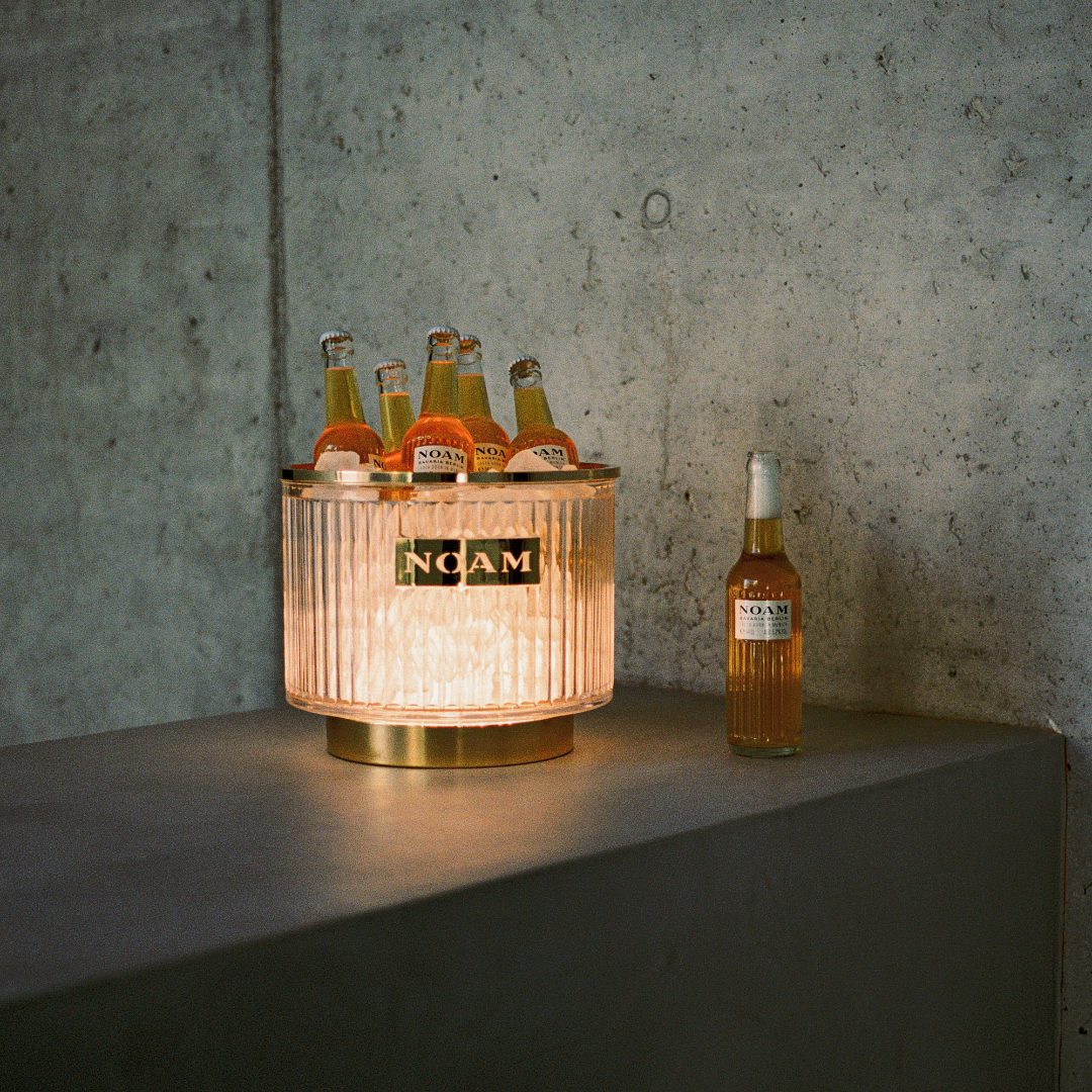 Gold ice bucket with bottles labeled 'Noma' against a concrete wall.