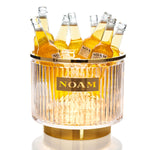A clear glass cooler with a gold base, labeled 'NOAM', filled with bottles, with an LED light illuminating the contents.