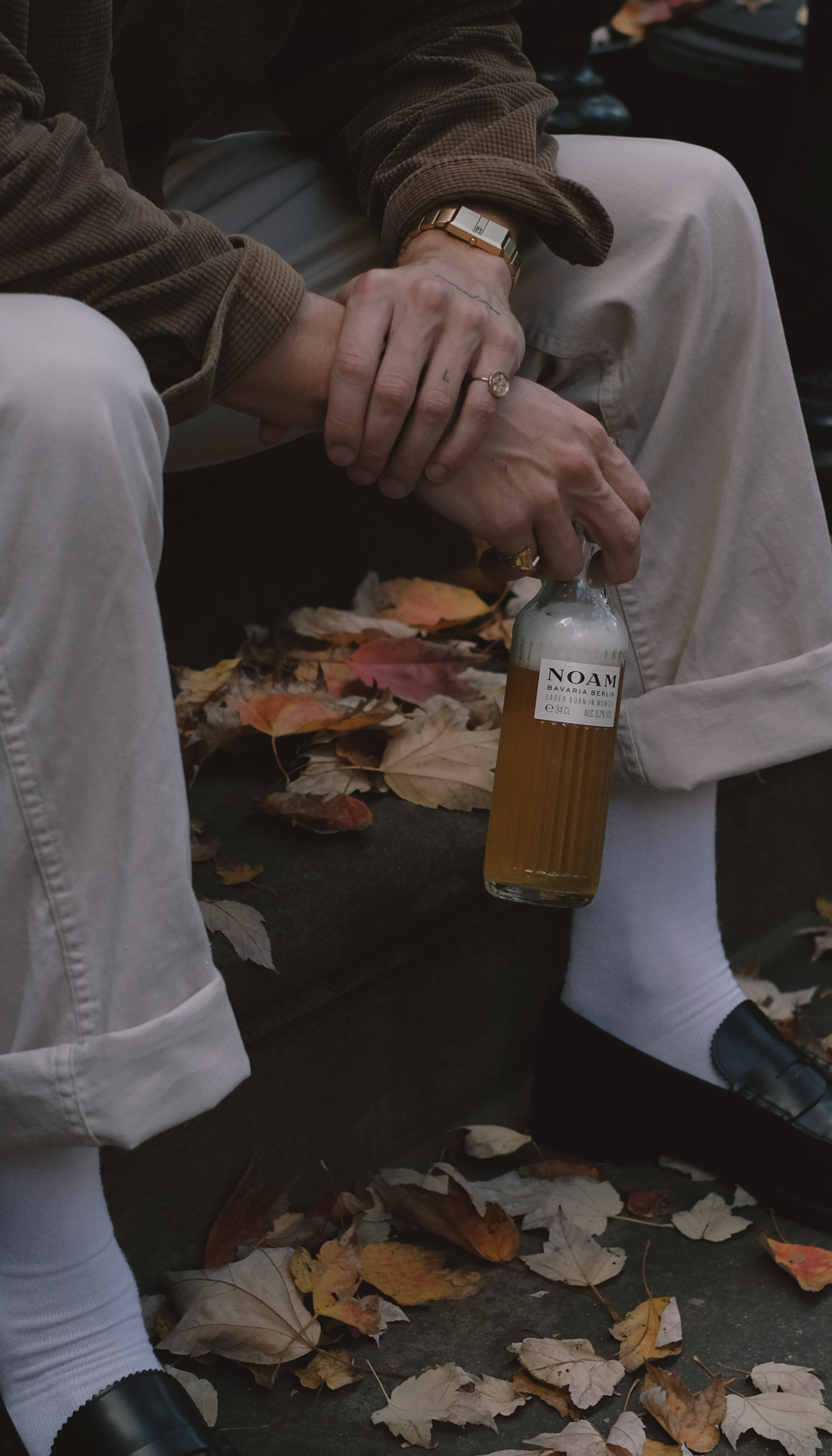 Person holding NOAM Beer amidst autumn leaves on steps.