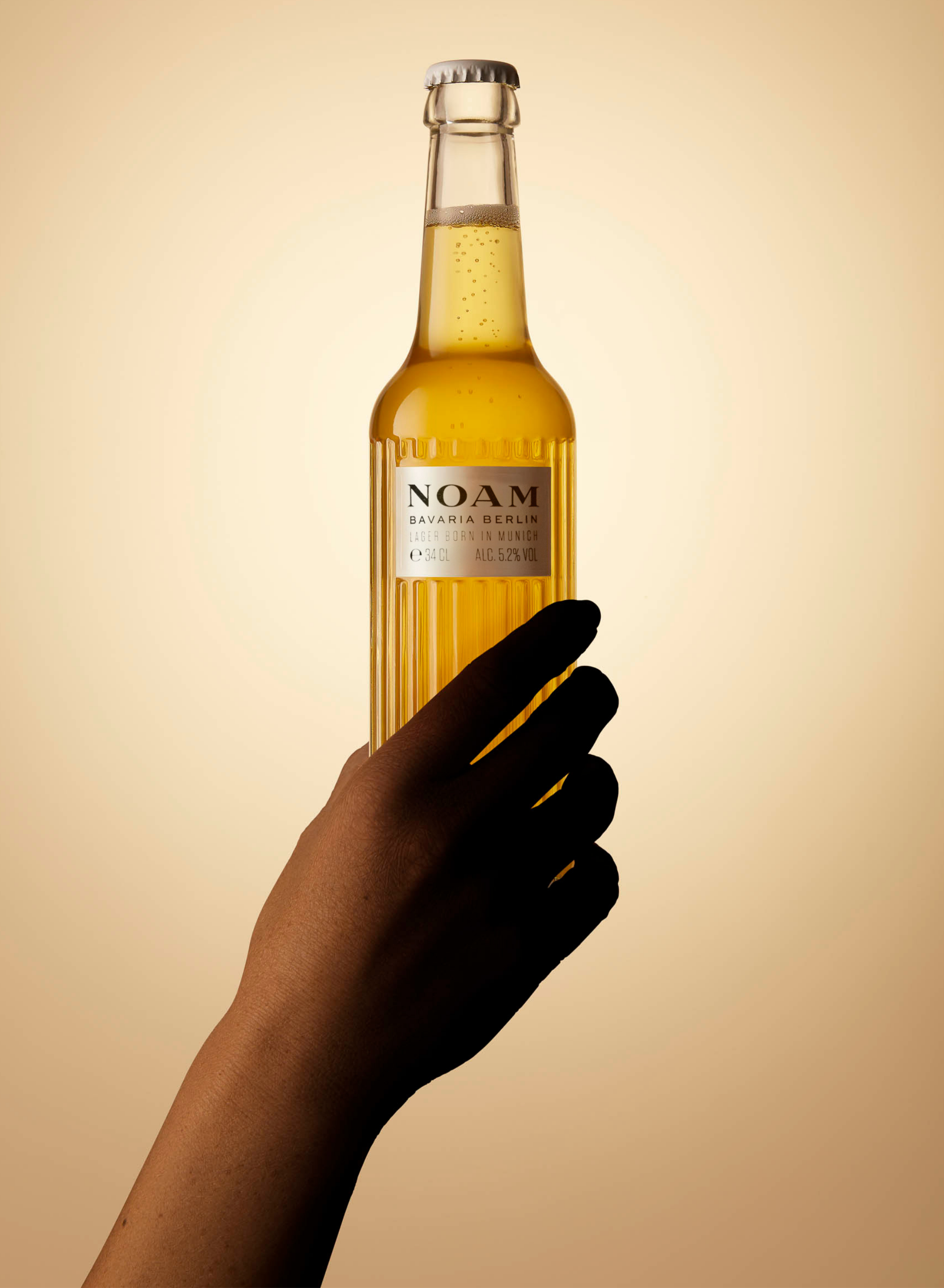 Hand holding a bottle of NOAM beer against a beige background - mobile image