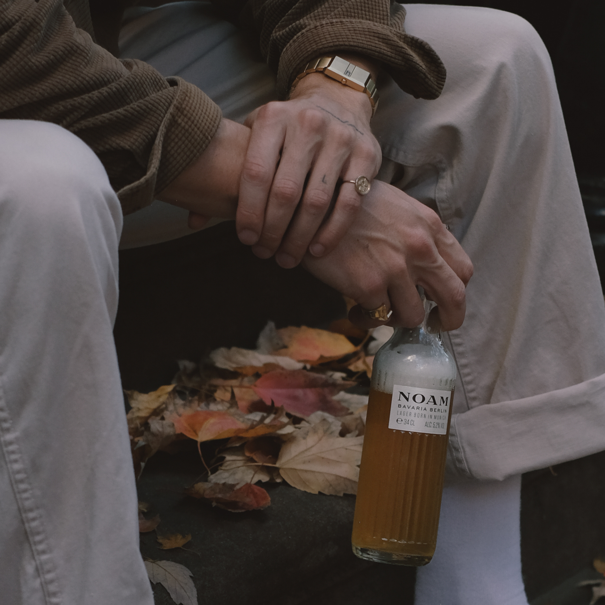 Person holding NOAM Beer amidst autumn leaves on steps.