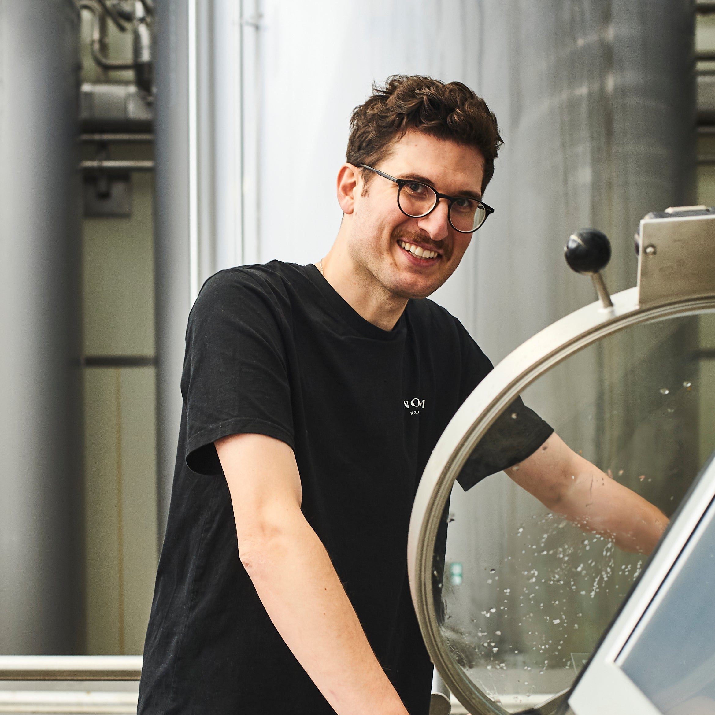Man wearing glasses and a black t-shirt standing next to a large round mirror with industrial background
