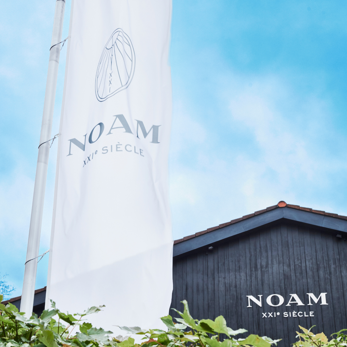 White sail with 'NOAM XXIe Siècle' branding against a blue sky, with a building featuring the same branding in the background.