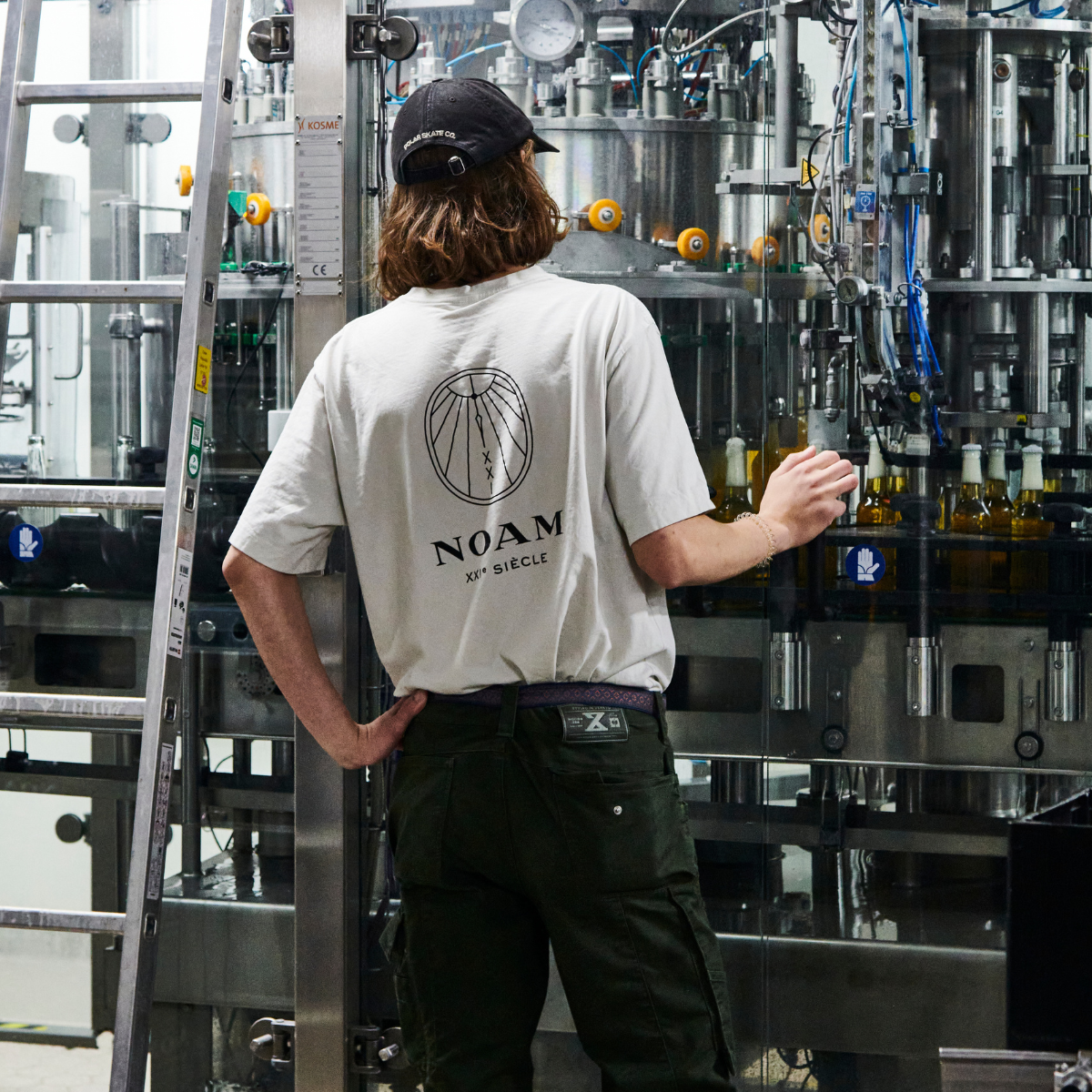 Person wearing a white t-shirt with 'NOAM' branding in an industrial setting